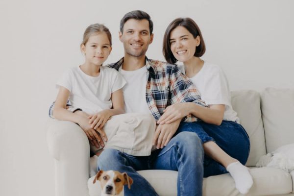 Horizontal view of three friendly family members and small pedigree dog pose at camera for making memorable photography. Dauther spends free time with mother and father, have good day at home